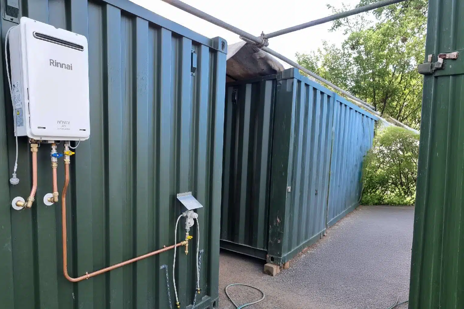 A detailed close-up shot of a white Rinnai Infinity external continuous flow gas hot water unit mounted on a corrugated dark green shipping container wall. The image clearly displays the unit's control panel, the Rinnai and Infinity branding, along with the detailed copper pipework, plumbing connections, and gas regulator, showing the installation on a site with other shipping containers and a gravel ground.